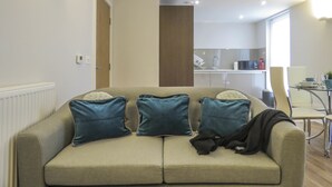 Een moderne woonkamer met een beige bank, teal kussens en een glazen salontafel. In de achtergrond is een keukenhoek zichtbaar met een spoelbak en kastjes.