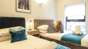 Een hotelkamer met twee bedden, een nachtkastje met een lamp en een raam met uitzicht op gebouwen.