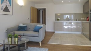 Een moderne woonkamer met een grijze bank, een glazen salontafel en een keuken met roestvrijstalen apparatuur.