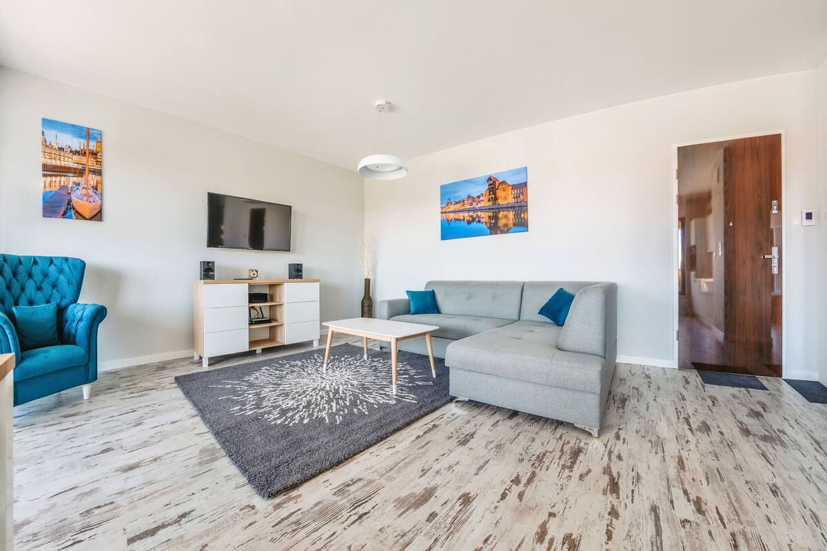 A modern living room with a grey sofa, a blue armchair, a wooden coffee table, and a grey rug. There are two paintings on the walls, a TV mounted above a wooden cabinet, and a wooden door.