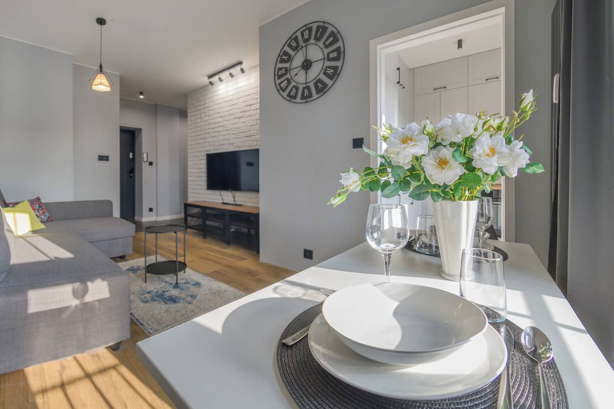 A modern living room with a grey sofa, a dining table set with plates and glasses, and a wall-mounted clock.