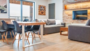 Un salon moderne comprenant un coin repas, un canapé gris, un téléviseur à écran plat fixé à un mur en briques et une grande fenêtre offrant une vue dégagée.