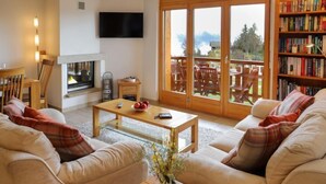 Una sala de estar acogedora con chimenea, televisor, estantería y un balcón con mobiliario exterior.