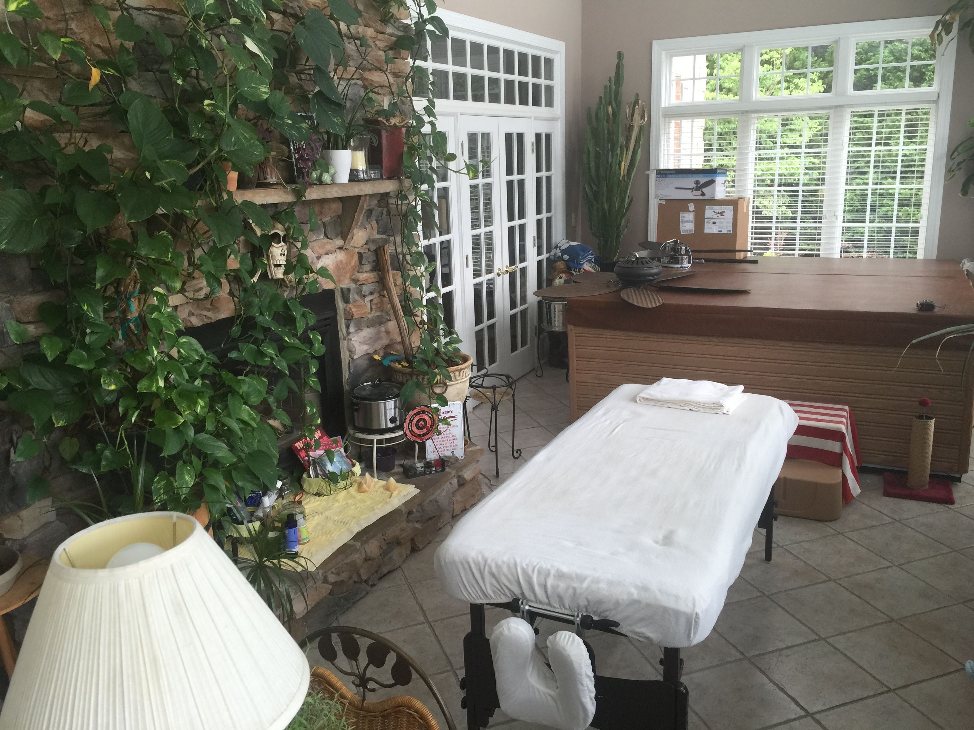 A cozy room with a massage table, a stone wall with greenery, a wooden counter, and a view through a window.