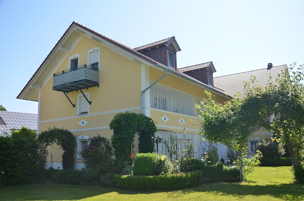 Una casa de dos pisos con balcón, rodeada de jardines cuidados y un cielo despejado.