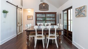A dining room with a wooden table and white chairs, a hanging light fixture, and a glass cabinet with dishes.
