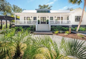A modern two-story house with a white exterior, a covered porch, and a landscaped garden with tropical plants.