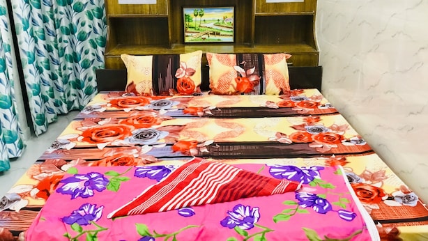 A bedroom with a bed, floral bedding, a shelf with decorative items, and curtains. - 2Bedroom Set Aastha @ GIP Mall Noida (Noida)