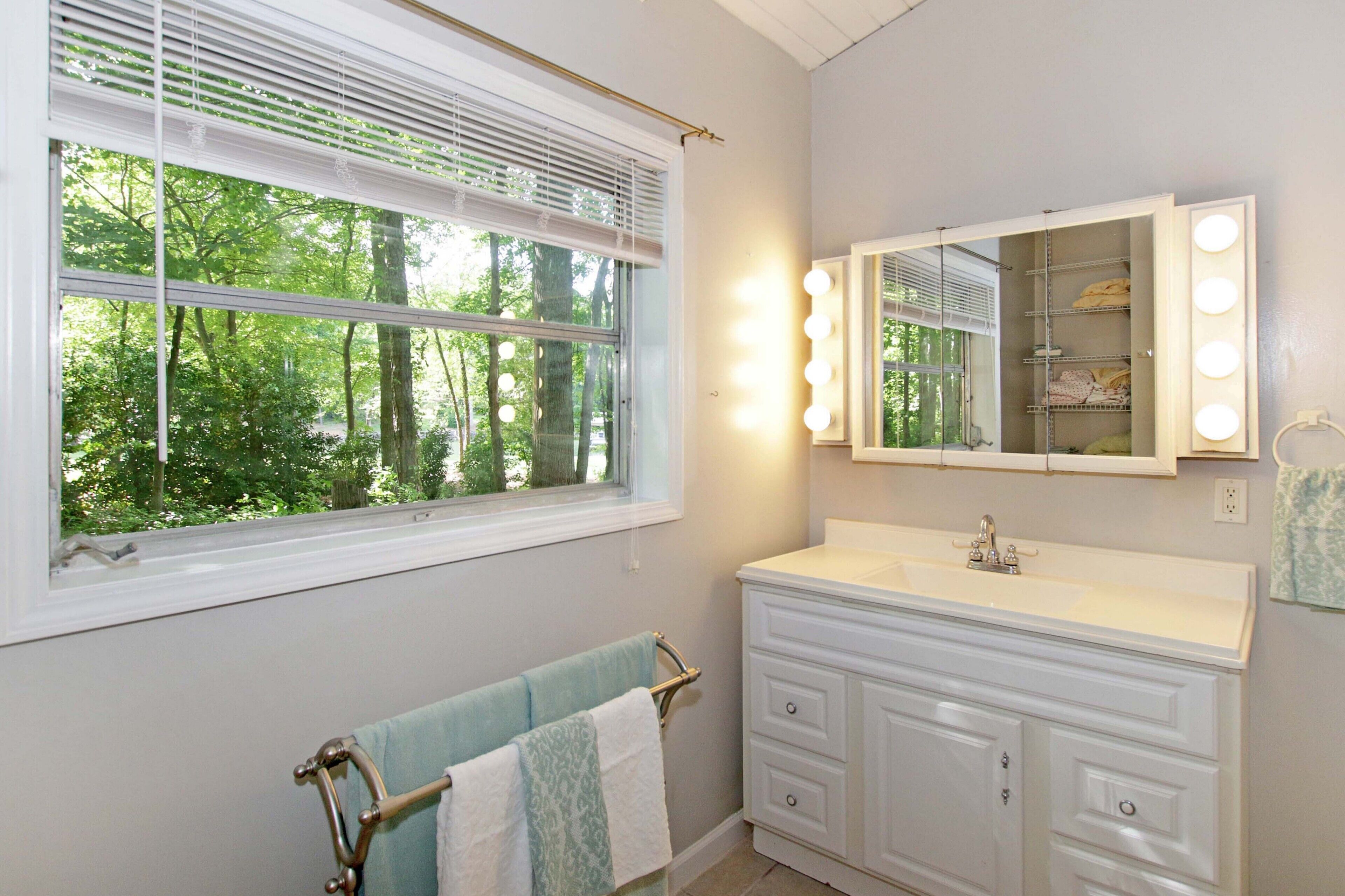 A bathroom with a vanity, a mirror with lights, a towel rack, and a window with blinds.