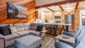 A cozy living area with a sofa, ottoman, and a coffee table, wooden cabinets, a kitchen with a refrigerator and microwave, and a painting of a mountain landscape. - June Lake Sierra Suns 4 -Spacious (June Lake)