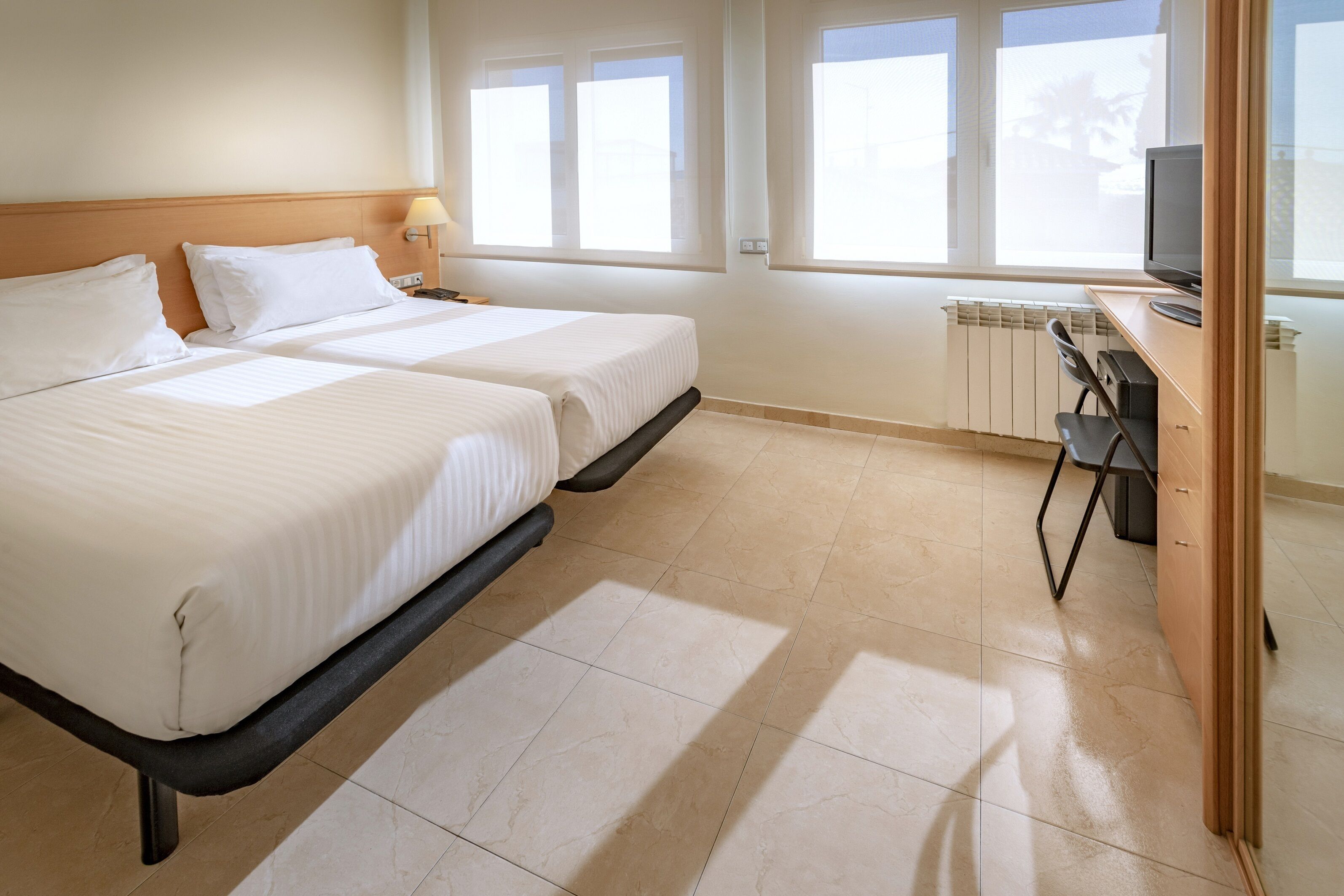 Una habitación de hotel con dos camas, un escritorio con una silla, televisión y ventanas amplias.