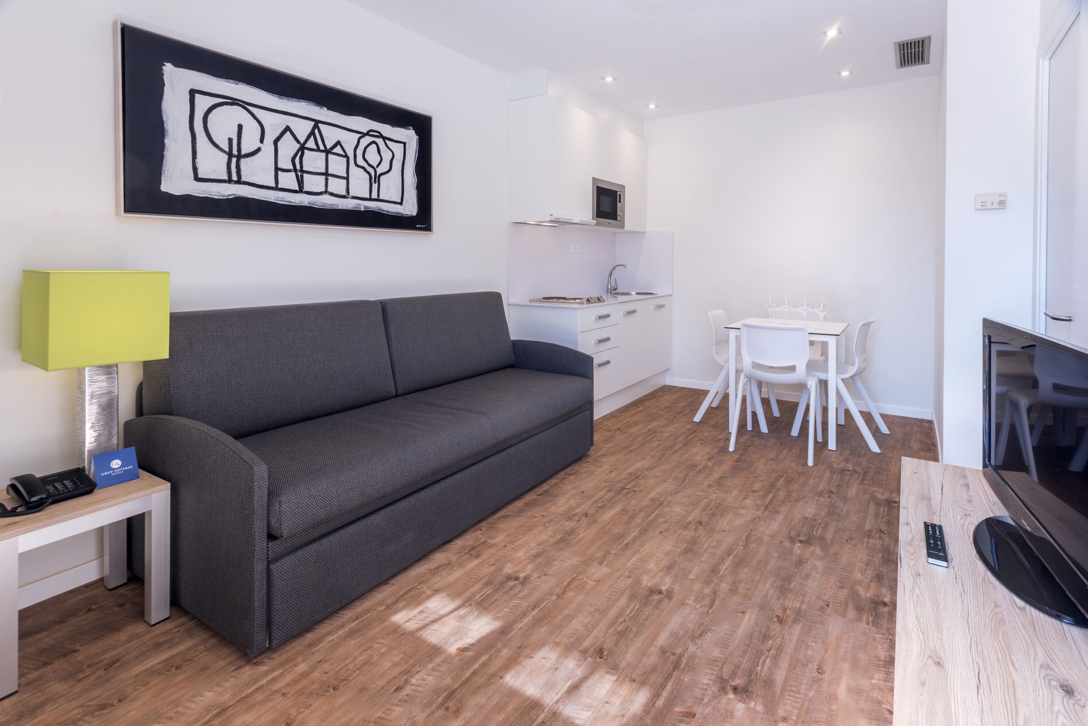 Una sala de estar moderna con un sofá gris, una mesita blanca, un televisor y una cocina al fondo.