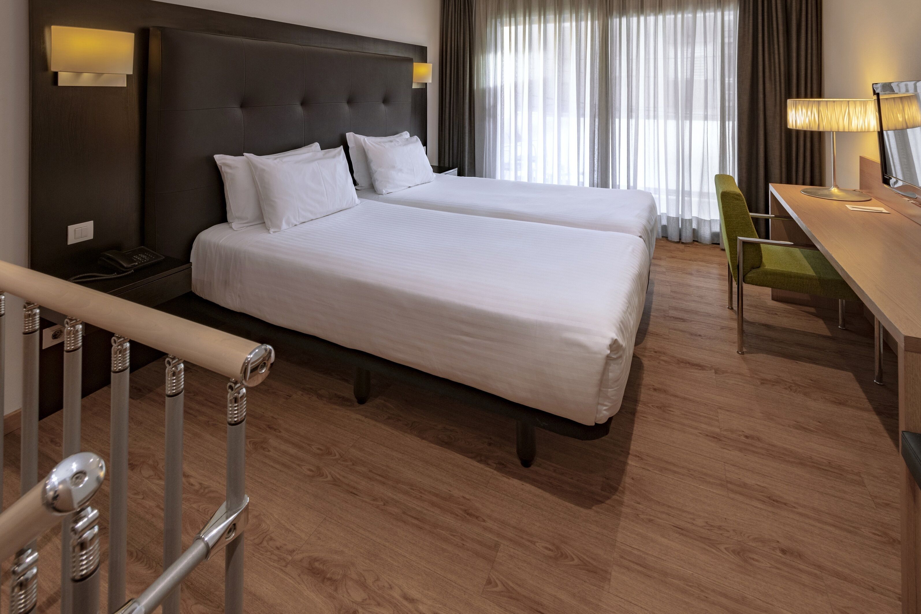 A hotel room with two beds, a desk, a chair, a lamp, and a view of the outdoors through large windows.