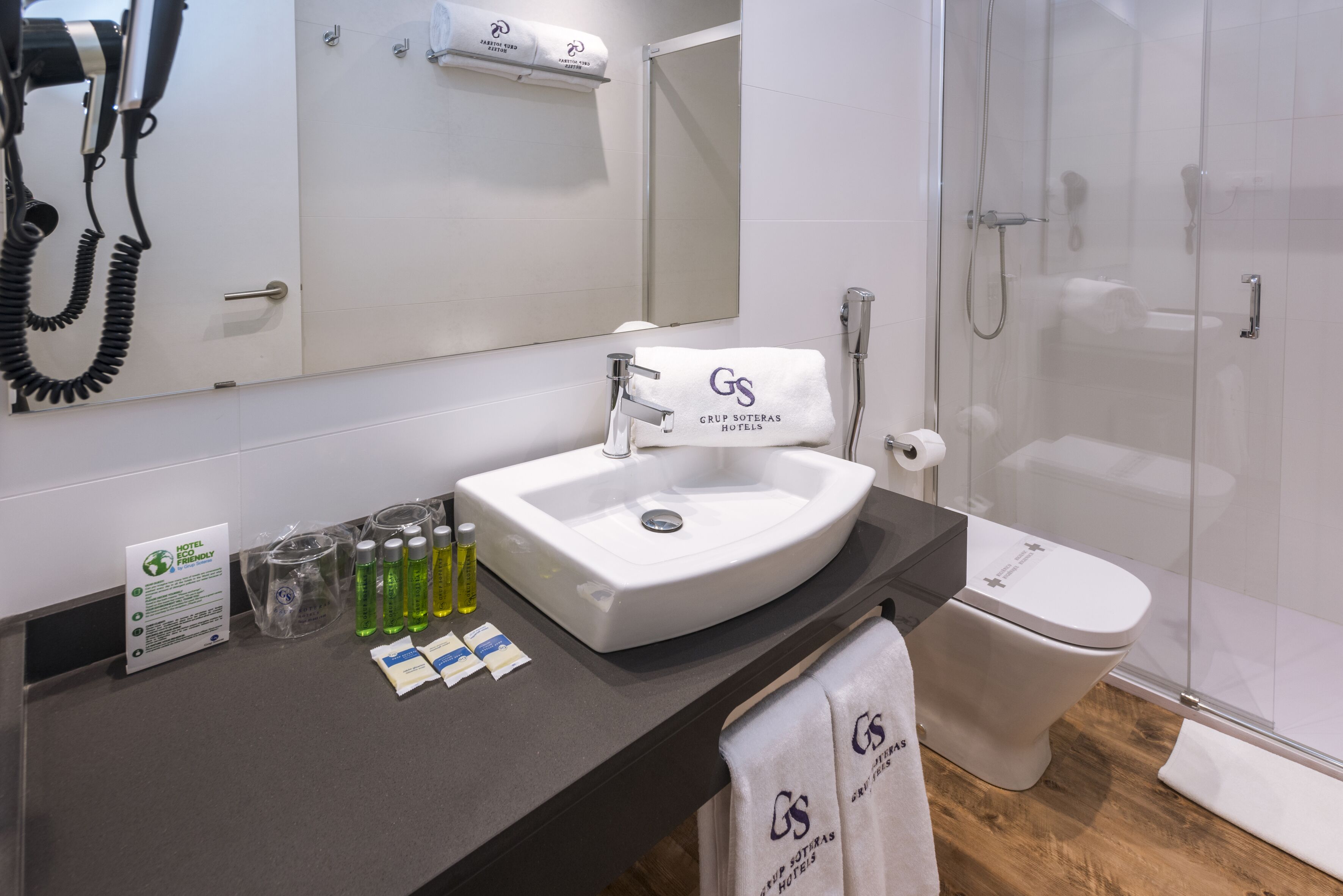 Et moderne badeværelse med en hvid vask, et spejl, en glasbrusekabine og et toilet. Der er håndklæder med hotellets logo, samt et skilt der angiver miljøvenlige tiltag.