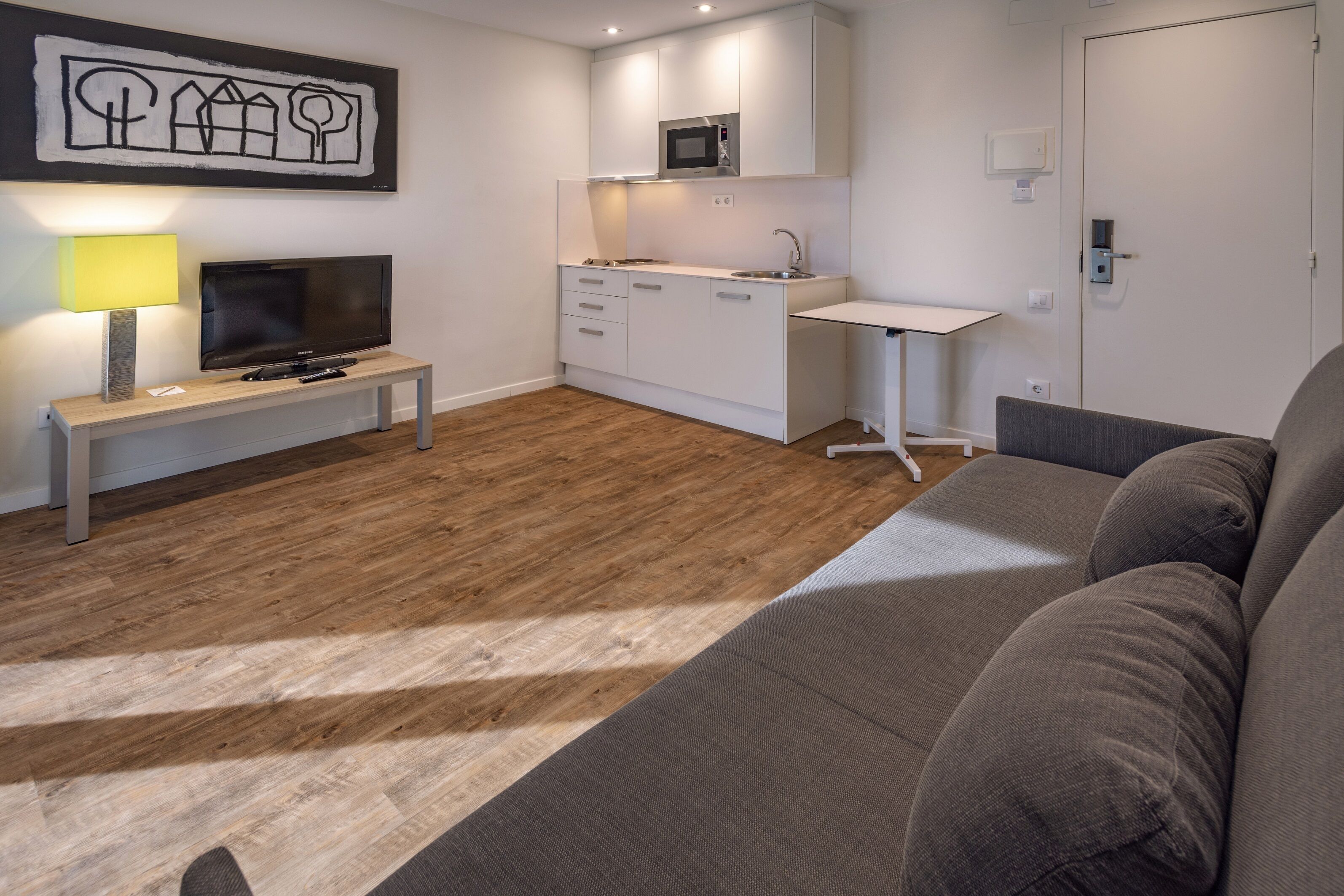 Una sala moderna con un sofá, un televisor, una mesita de noche con una lámpara, y una zona de cocina con armarios y un microondas.