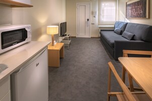 Um espaço compacto com cozinha compacta, incluindo micro-ondas, frigorífico e uma pequena mesa com cadeiras. Há um sofá e uma televisão sobre um suporte.
