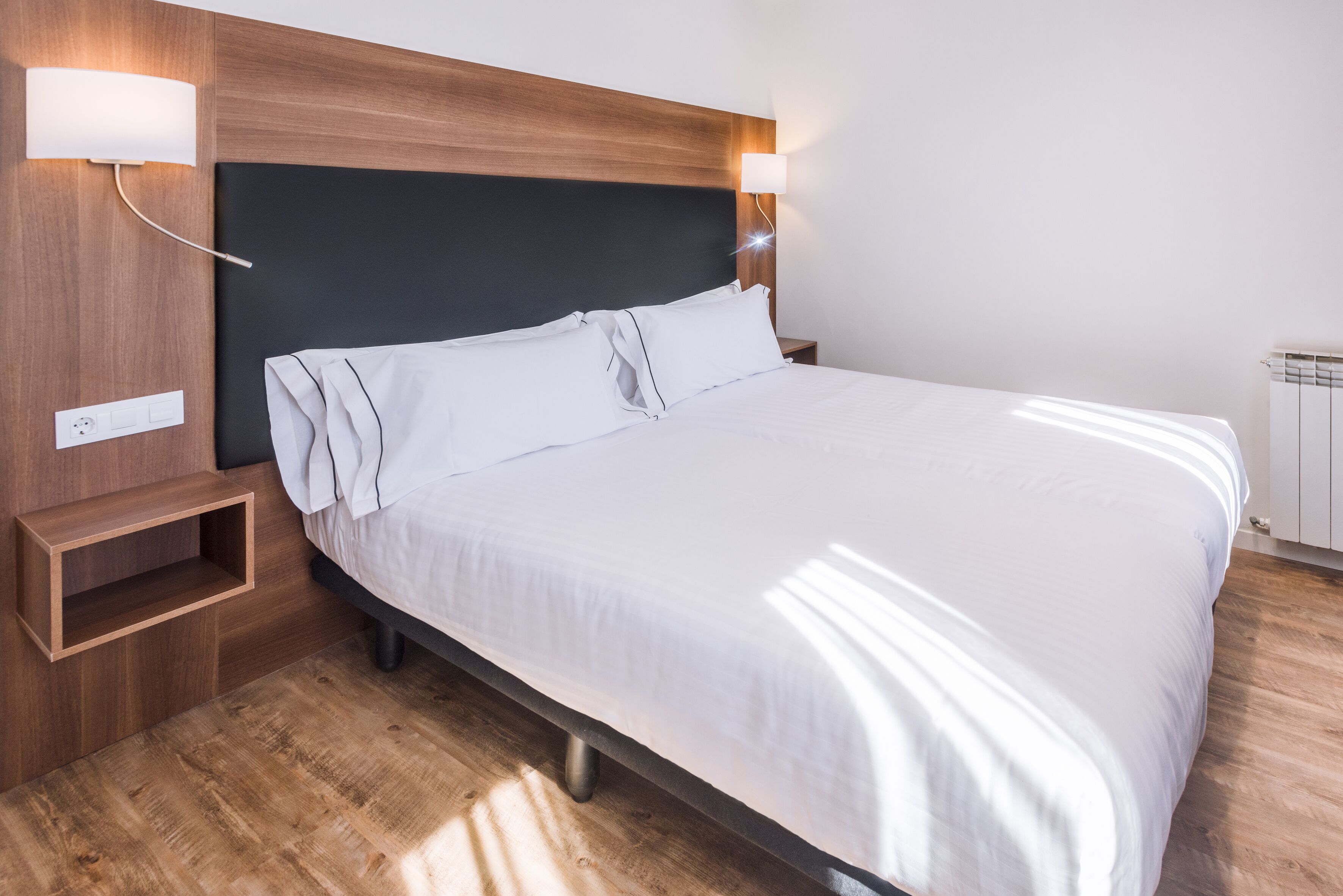 Una cama bien hecha con sábanas blancas, cabecera de madera y lámparas de noche.