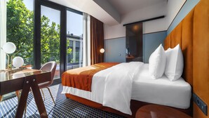 Habitación de hotel con una cama grande, un escritorio con silla, un espejo y vistas a árboles y edificios a través de amplias ventanas.