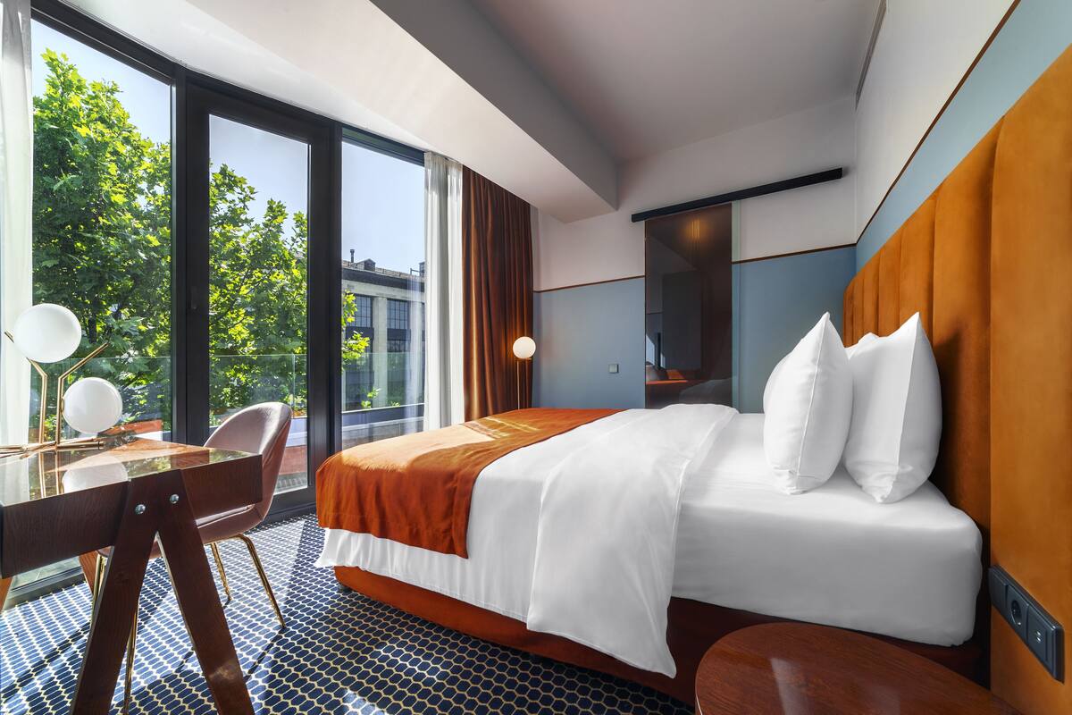 A hotel room with a large bed, a desk with a chair, a mirror, and a view of trees and buildings through large windows.
