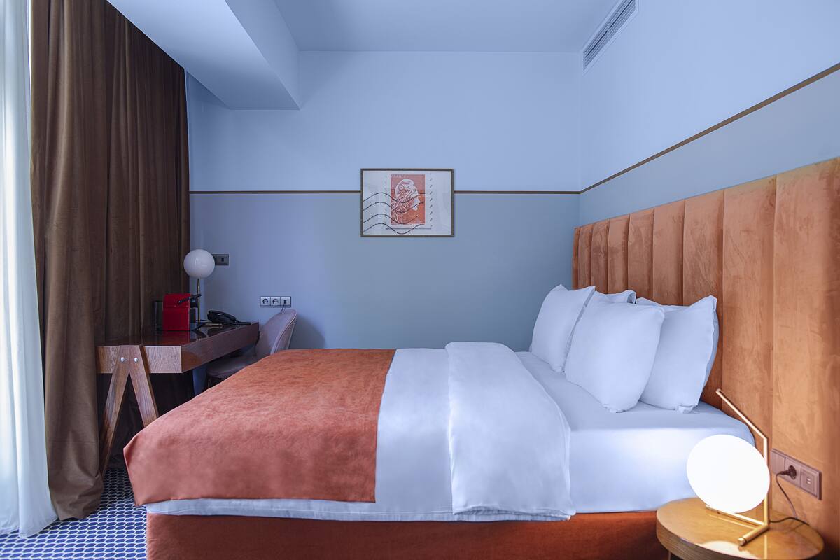 A hotel room with a bed, a desk, a chair, a lamp, and a framed picture on the wall.