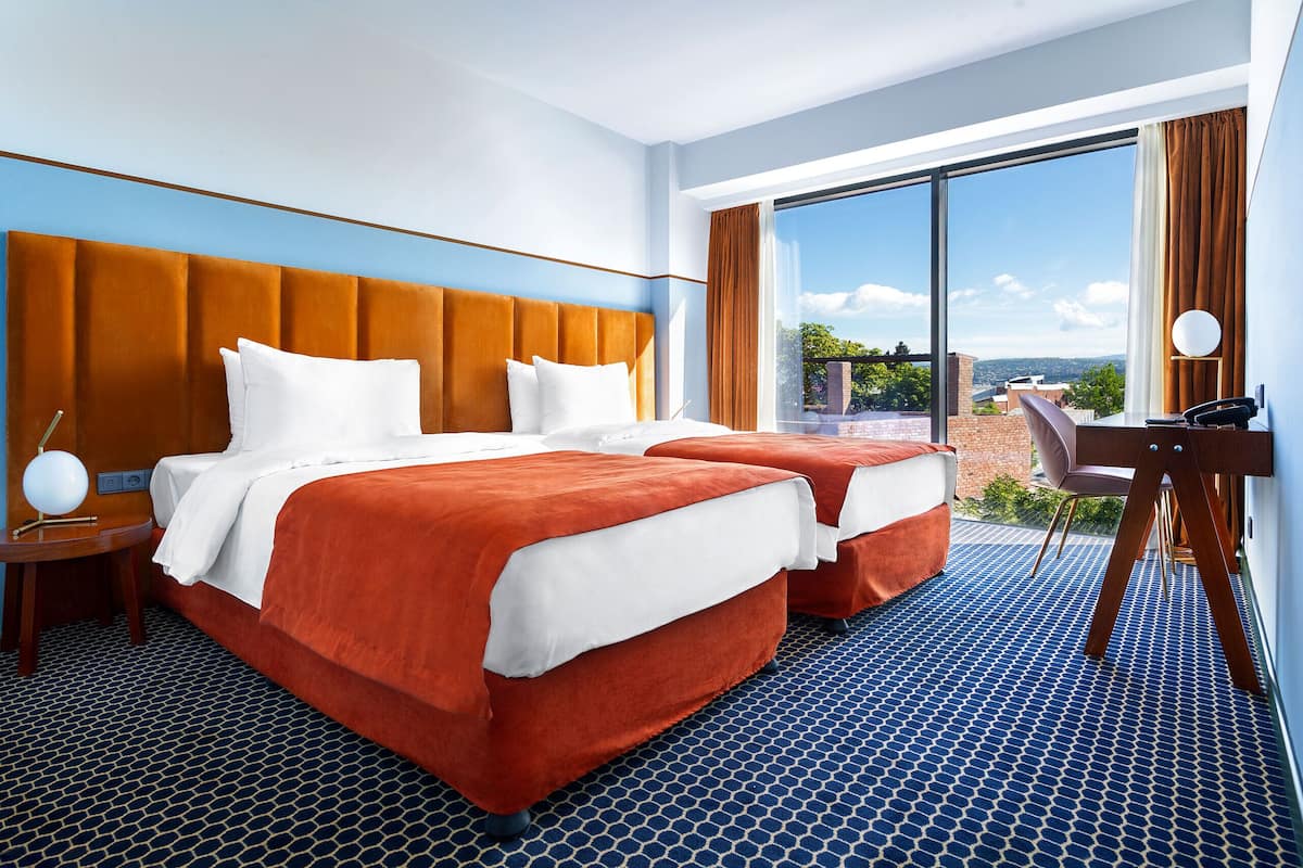 A hotel room with two beds, a desk, and a large window offering a city view.