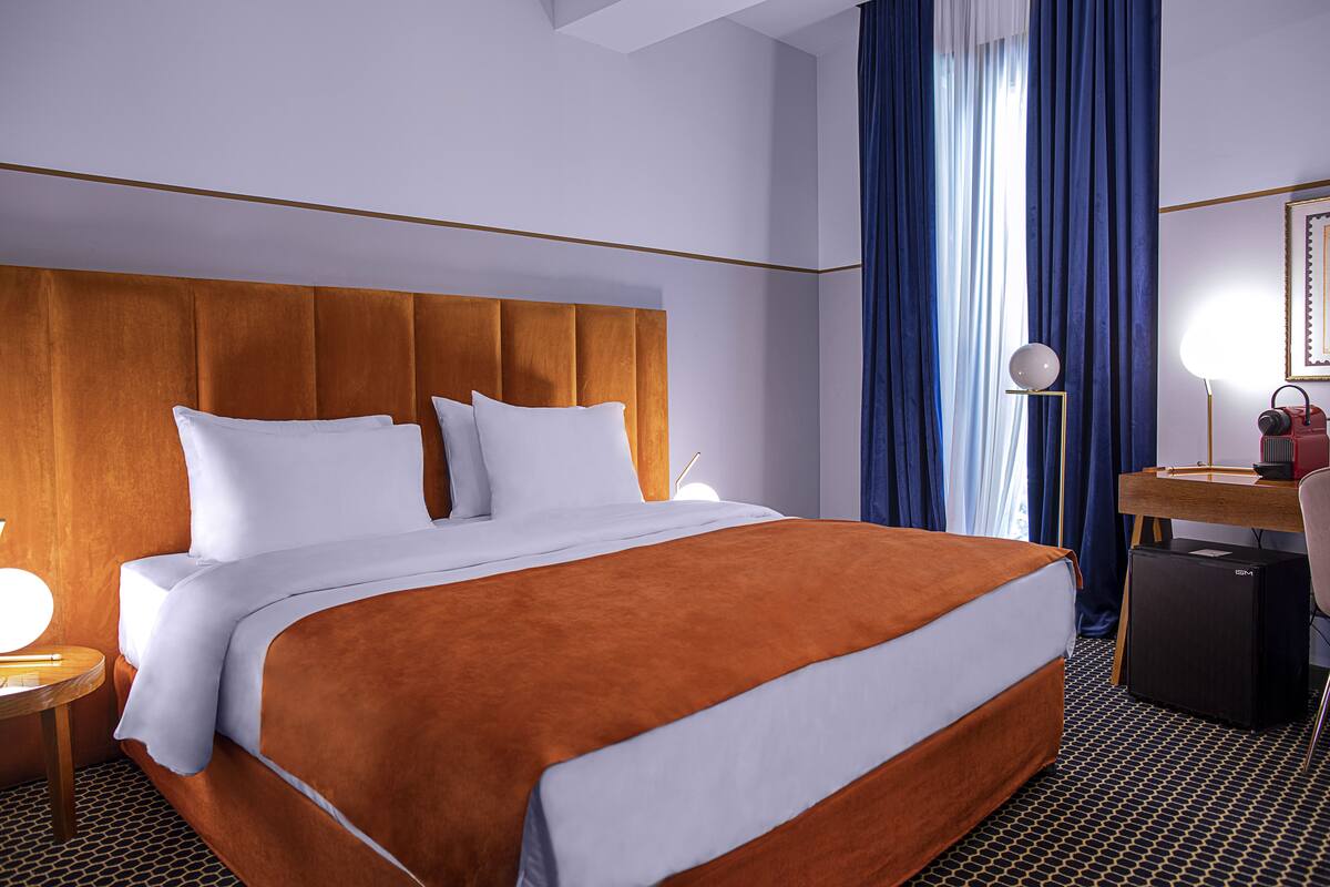 A hotel room with a large bed, a desk with a lamp, and a compact refrigerator.