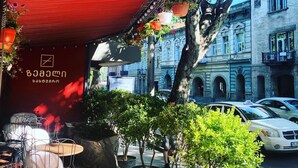 Un toldo rojo con luces colgantes y plantas en macetas, un letrero con un logotipo y un árbol al fondo.
