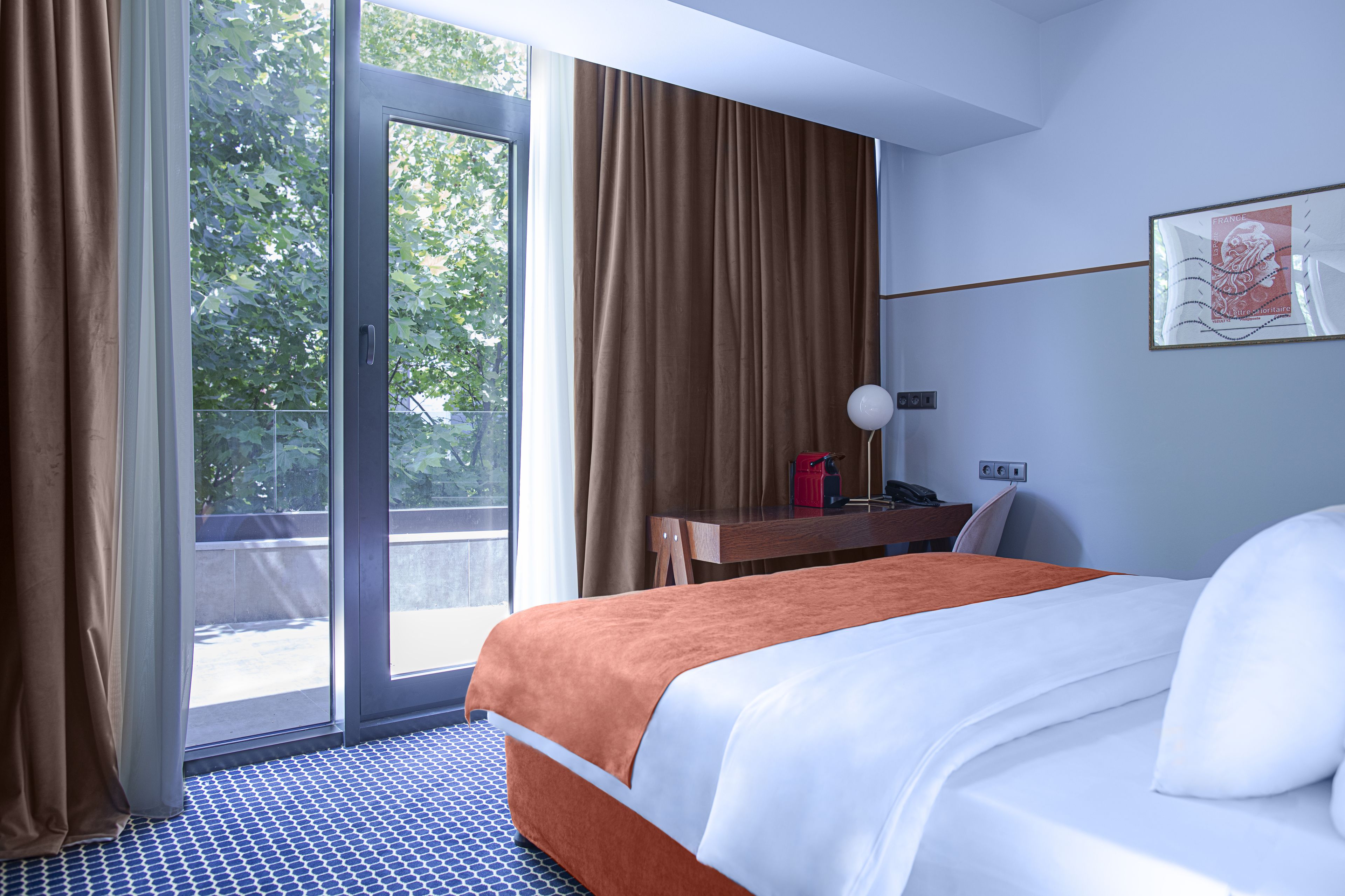 Een hotelkamer met een bed, een bureau, een stoel, een lamp en een raam met gordijnen.