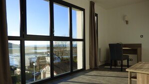 A room with large windows offering a view of a body of water and buildings, a wooden desk, a chair, and a radiator on the wall. - Hôtel Les Galets (Criel-sur-Mer)