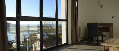 A room with large windows offering a view of a body of water and buildings, a wooden desk, a chair, and a radiator on the wall.