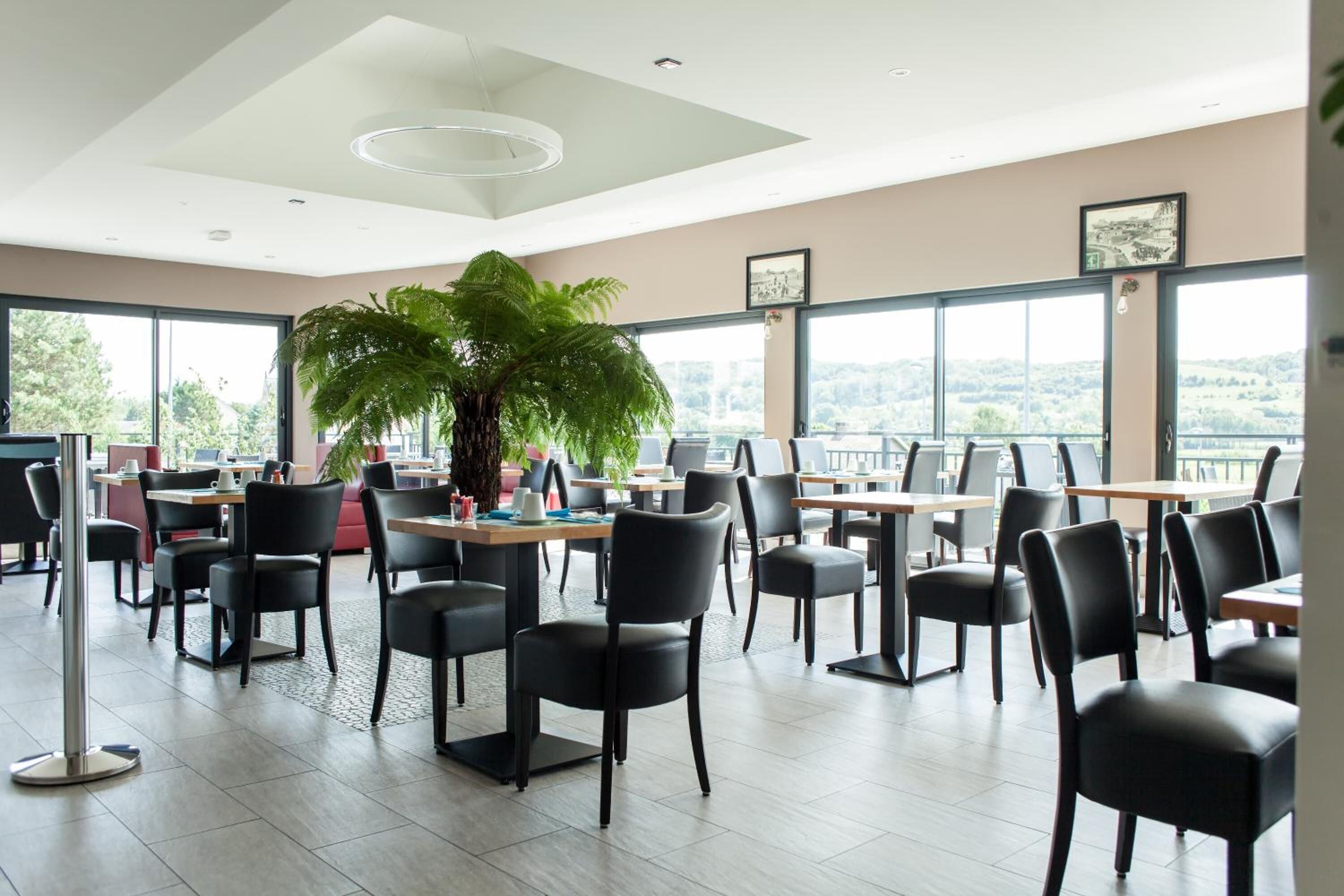 Un comedor moderno con mesas y sillas, ventanas amplias y una planta en el centro.