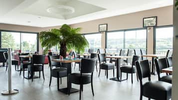 Un espace repas moderne, doté de tables et de chaises, de grandes fenêtres et d’une plante au centre.