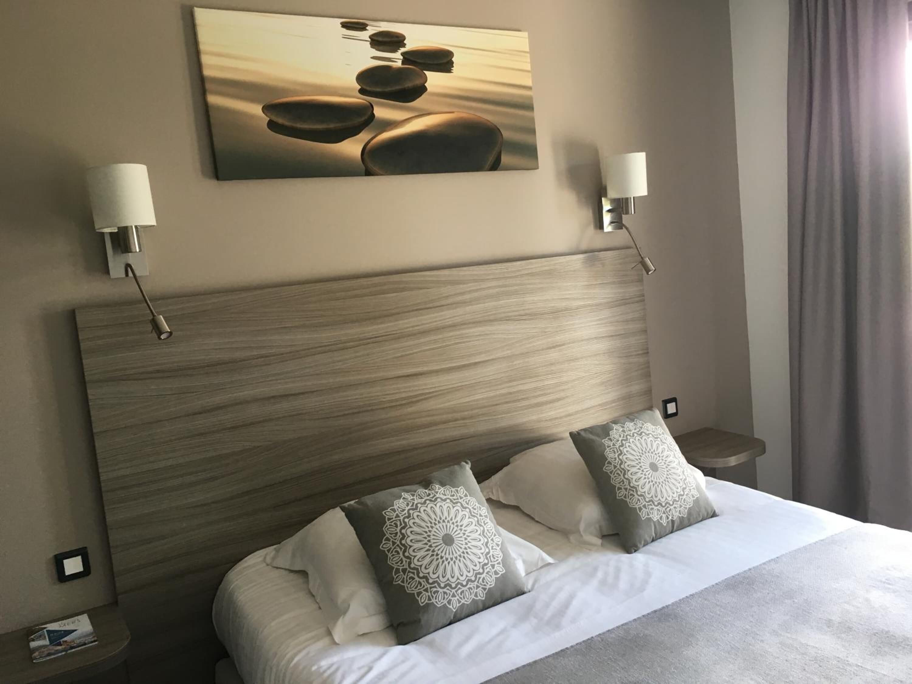 A hotel room with a wooden headboard, two bedside lamps, a bed with white bedding and patterned pillows, a painting of rocks on the wall, and a window with curtains.