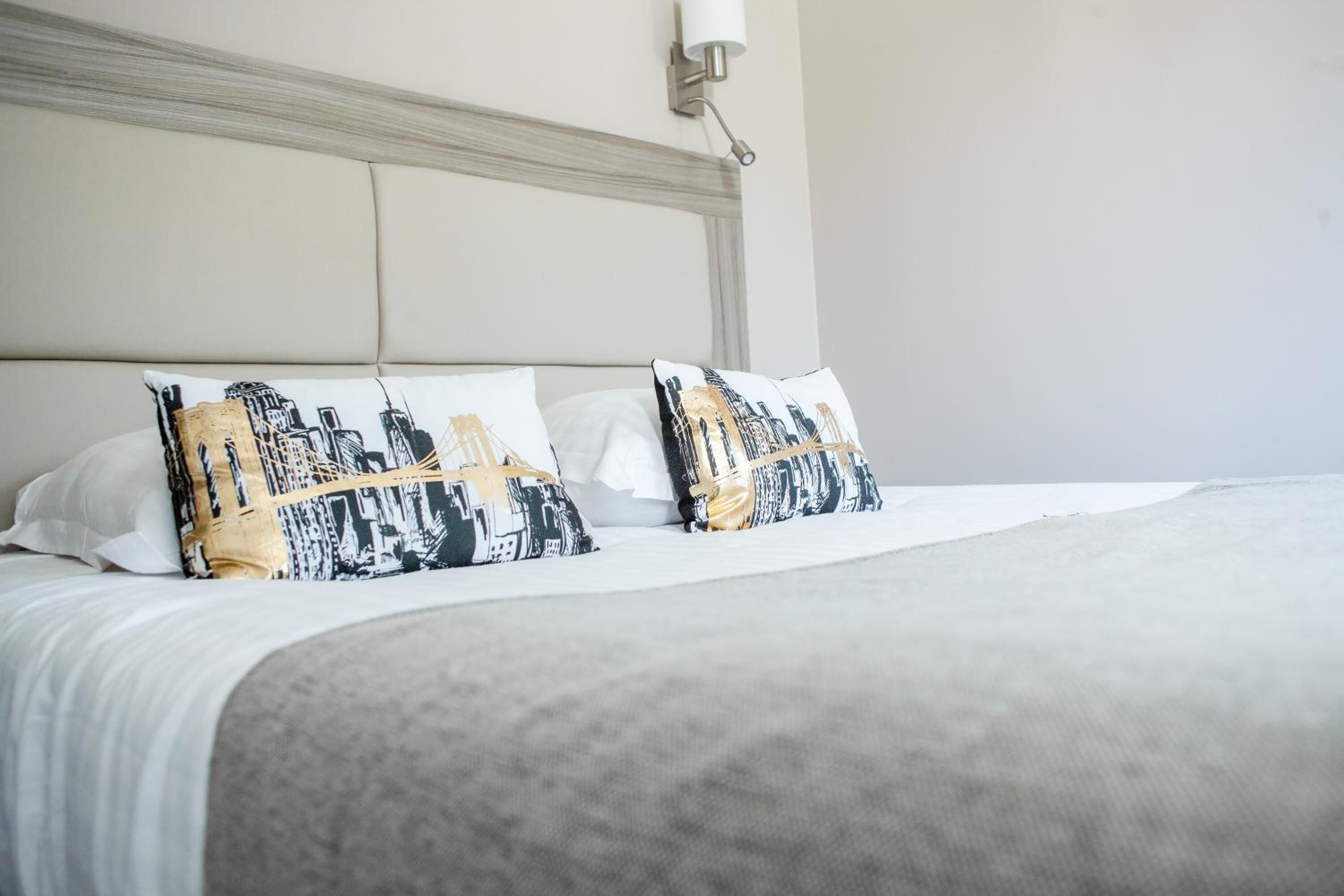 Ein ordentlich bezogenes Bett mit zwei dekorativen Kissen mit Stadtansicht-Motiv, einem Kopfteil mit einer Leuchte und einer schlichten Wand.
