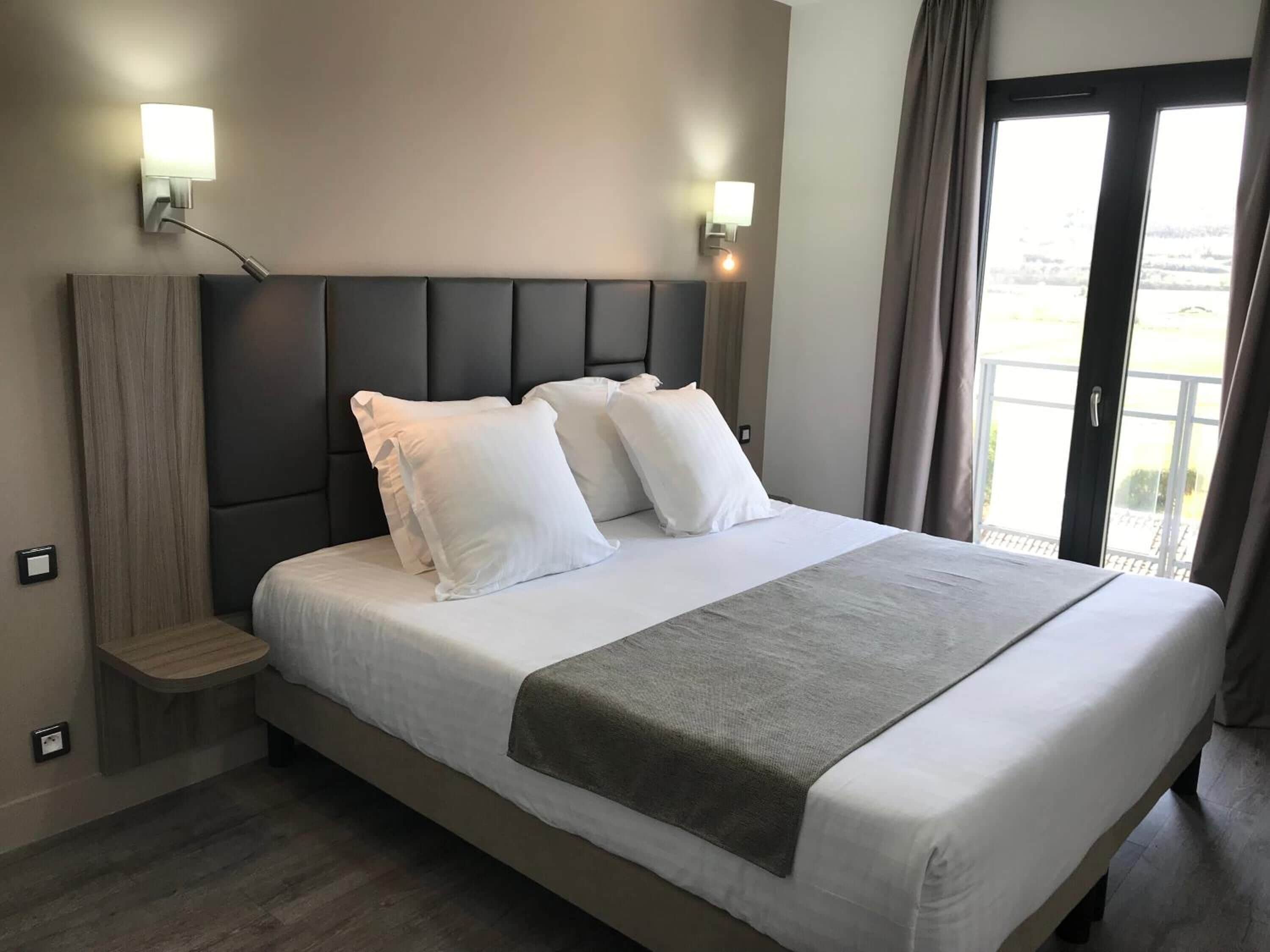A hotel room with a large bed, two bedside lamps, a small wooden nightstand, and a view of the outdoors through a sliding door.