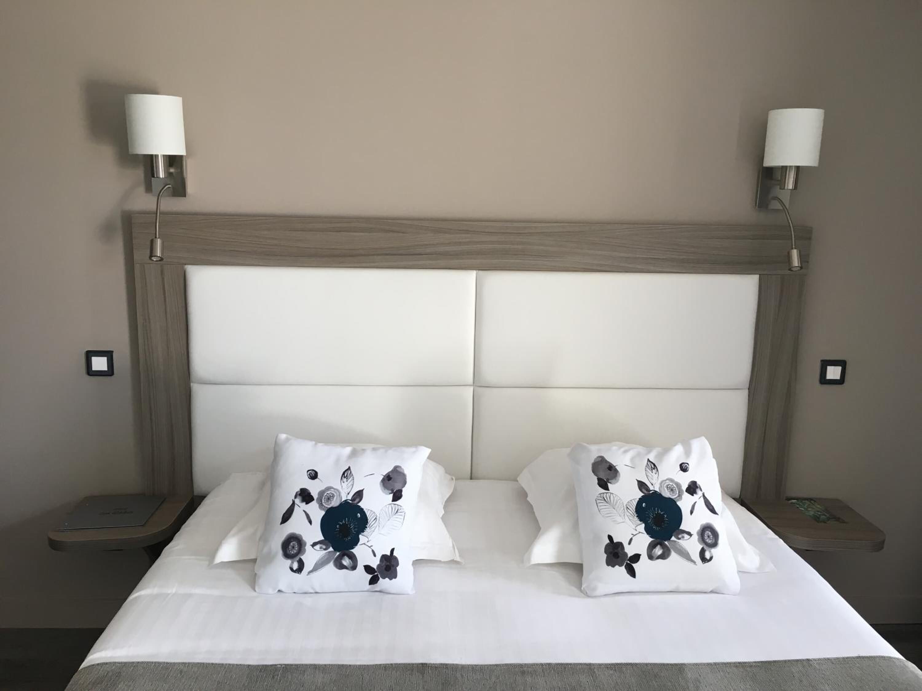 Een bed met een houten hoofdbord, twee witte kussens met bloemendesign en twee bedlampjes.
