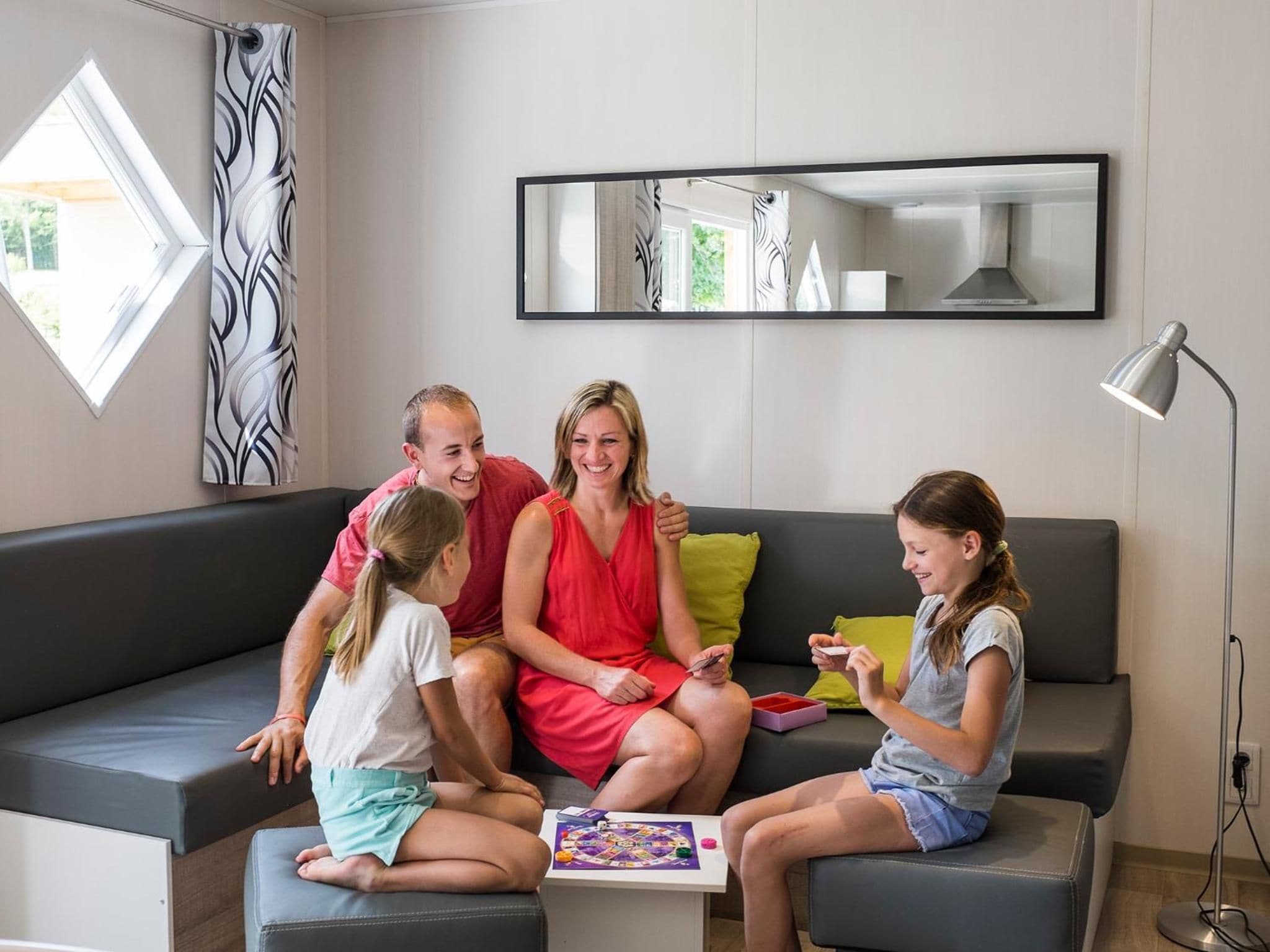 Una familia de cuatro personas en un sofá moderno gris con una pequeña mesa delantera, una ventana grande con cortinas y una lámpara de pie.