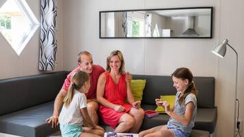 Una familia de cuatro personas en un sofá moderno gris con una pequeña mesa delantera, una ventana grande con cortinas y una lámpara de pie.