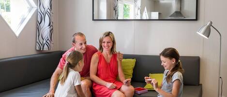 A family of four on a modern grey sofa with a small table in front, a large window with curtains, and a floor lamp.
