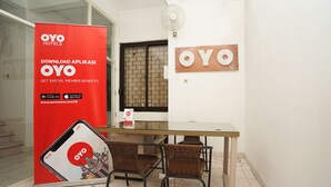 Uma área de recepção de hotel com um banner promocional vermelho, uma placa de madeira, uma mesa de vidro, cadeiras de vime e uma janela com vista.
