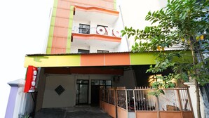 Um edifício de vários andares com fachada verde e laranja, entrada com portão e uma pequena árvore em frente.