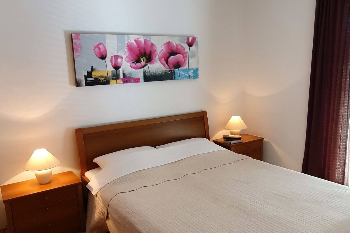 A bedroom with a wooden bed, two bedside tables with lamps, a painting of flowers on the wall, and a window with curtains.