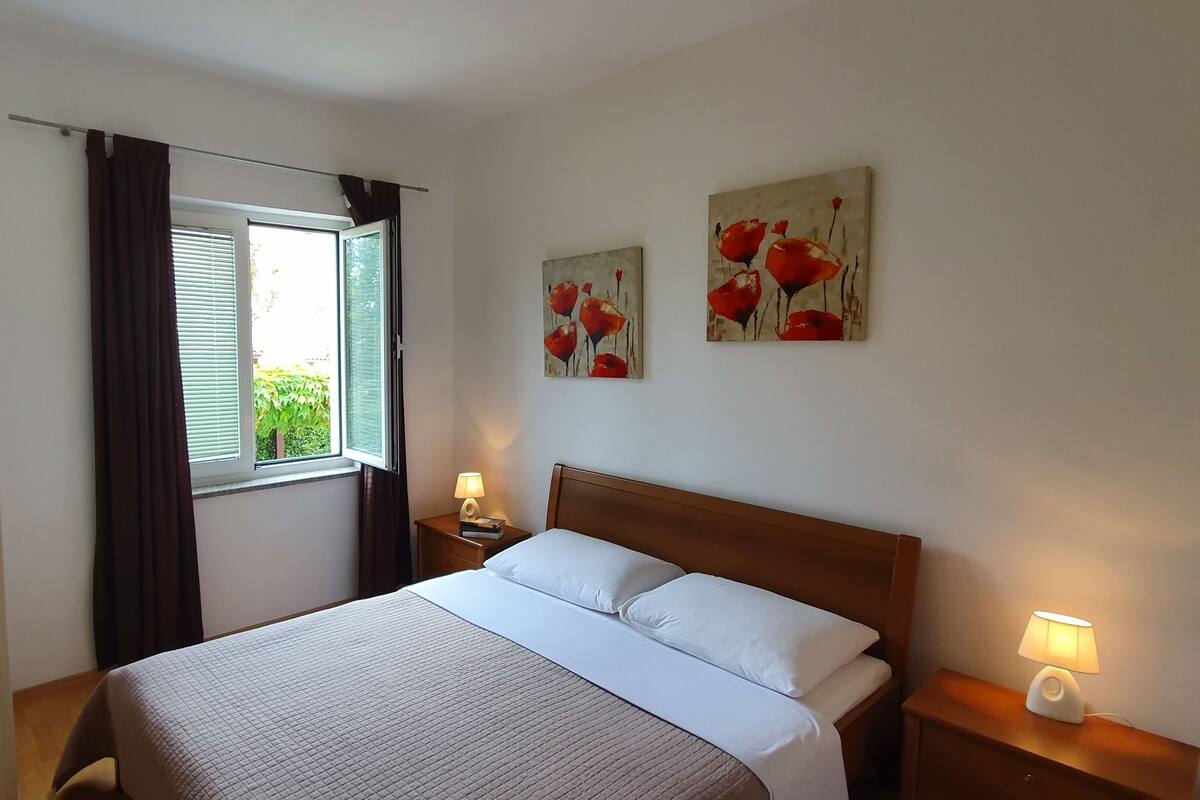 A hotel room with a bed, a nightstand with a lamp, a window with curtains, and two wall art pieces.
