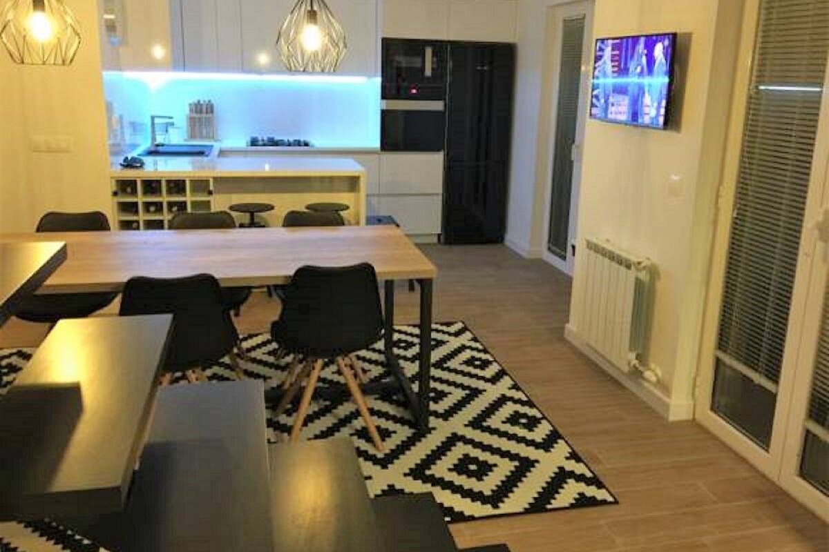 A modern kitchen with white cabinets, a black refrigerator, and a built-in oven. There is a dining area with a wooden table and chairs, and a black and white patterned rug.