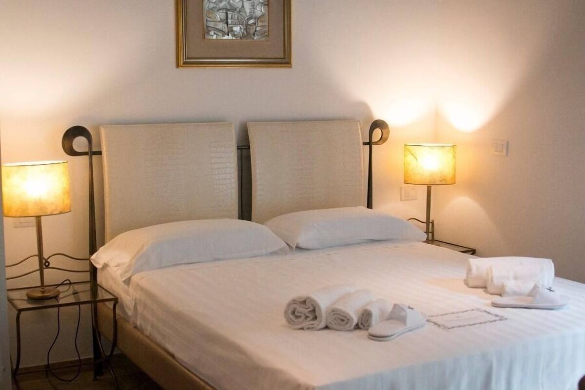 A neatly made bed with white linens and rolled towels, two bedside lamps, and a framed artwork on the wall.