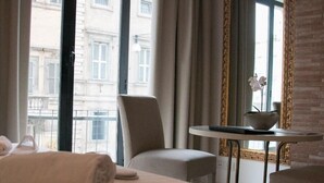 Quarto de hotel com cama, uma cadeira, uma mesinha com uma tigela e janela com cortinas.