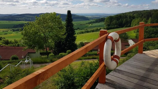 Une terrasse en bois, agrémentée d’une bouée de sauvetage, qui offre une vue sur un paysage verdoyant, avec des arbres et des collines à l’horizon.