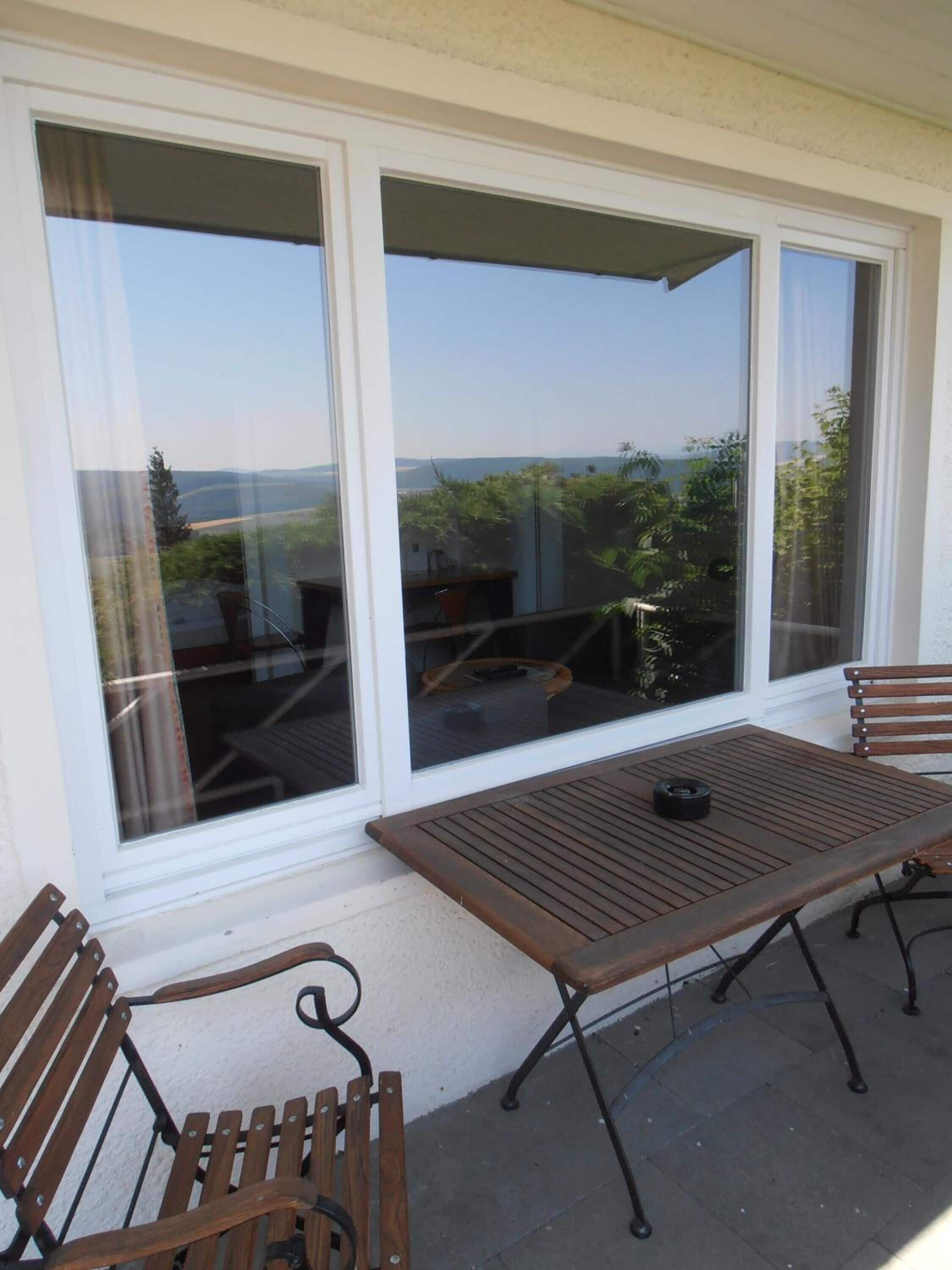 Un patio con una mesa y dos sillas de madera, ventanas amplias con vistas a zonas verdes y un paisaje lejano.
