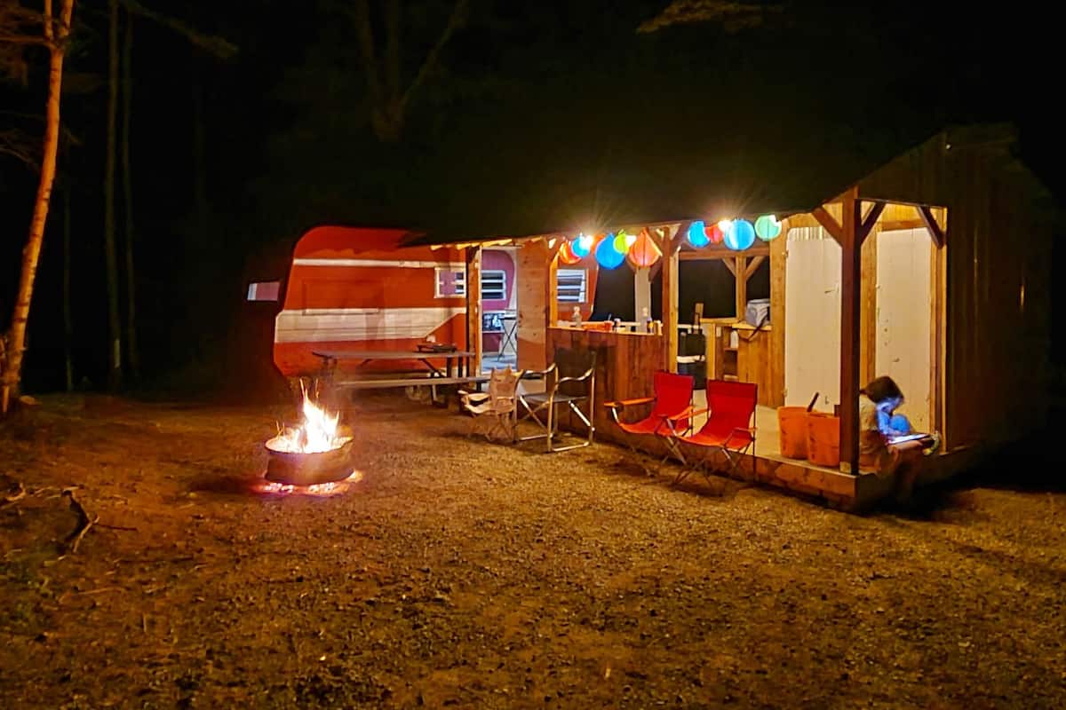 A rustic campsite with a fire, a wooden structure, and a person working on a laptop.