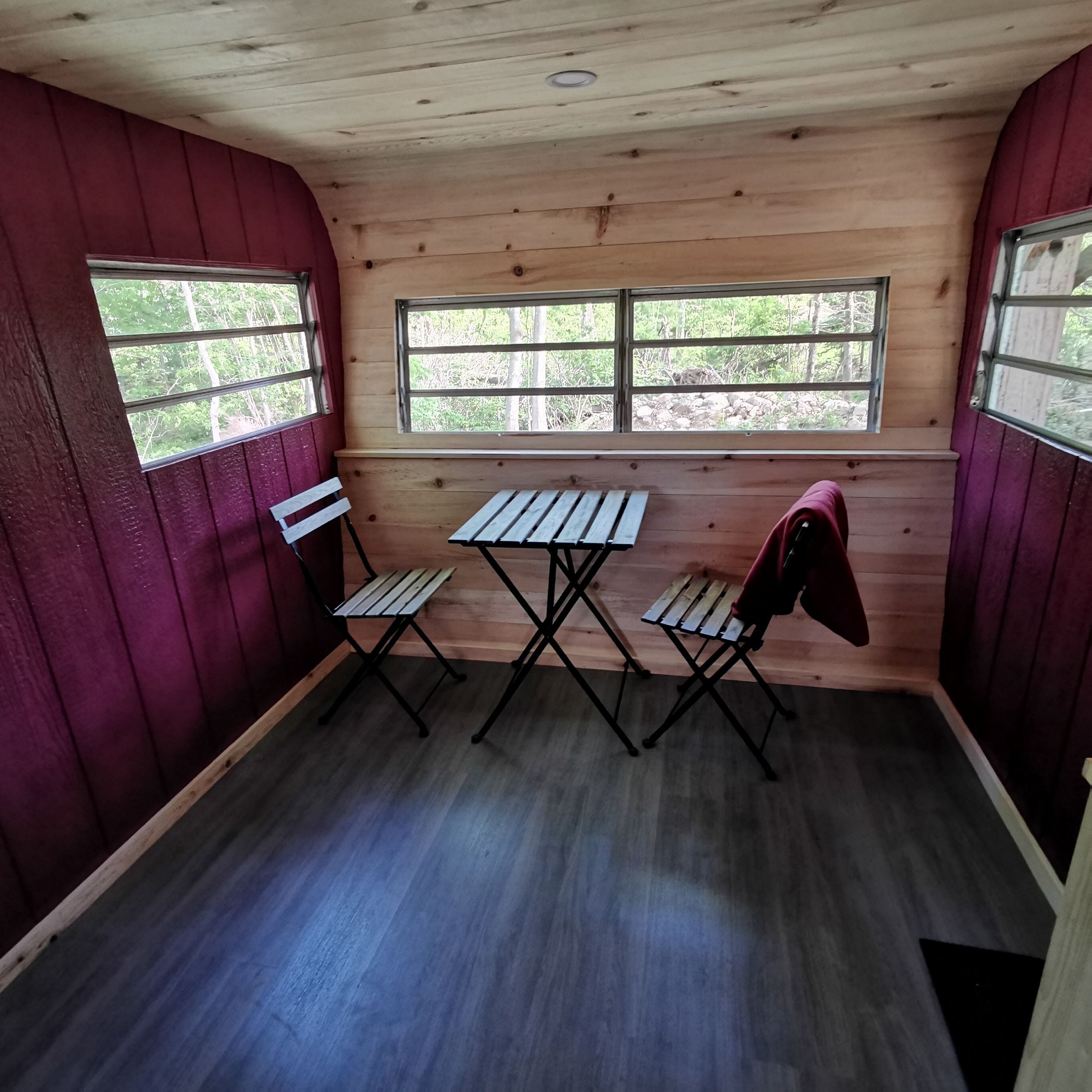 A small room with wooden walls and a sloped ceiling. There are two windows with metal bars, a wooden bench, and a folding table.